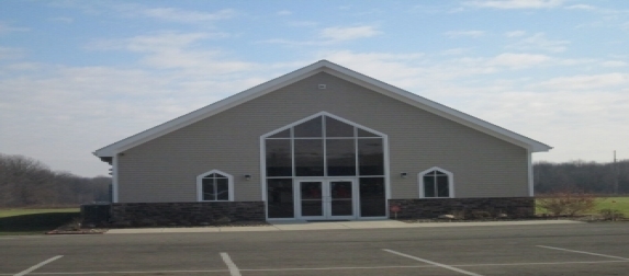 The church building: a gray building with a tall arched window above glass front doors